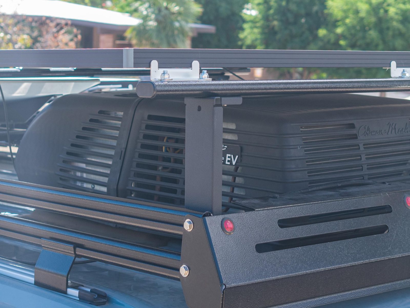 Rear corner of a van roof rack with a Desert Vans Mesa Riser holding a crossbar and solar panel over the A/C unit